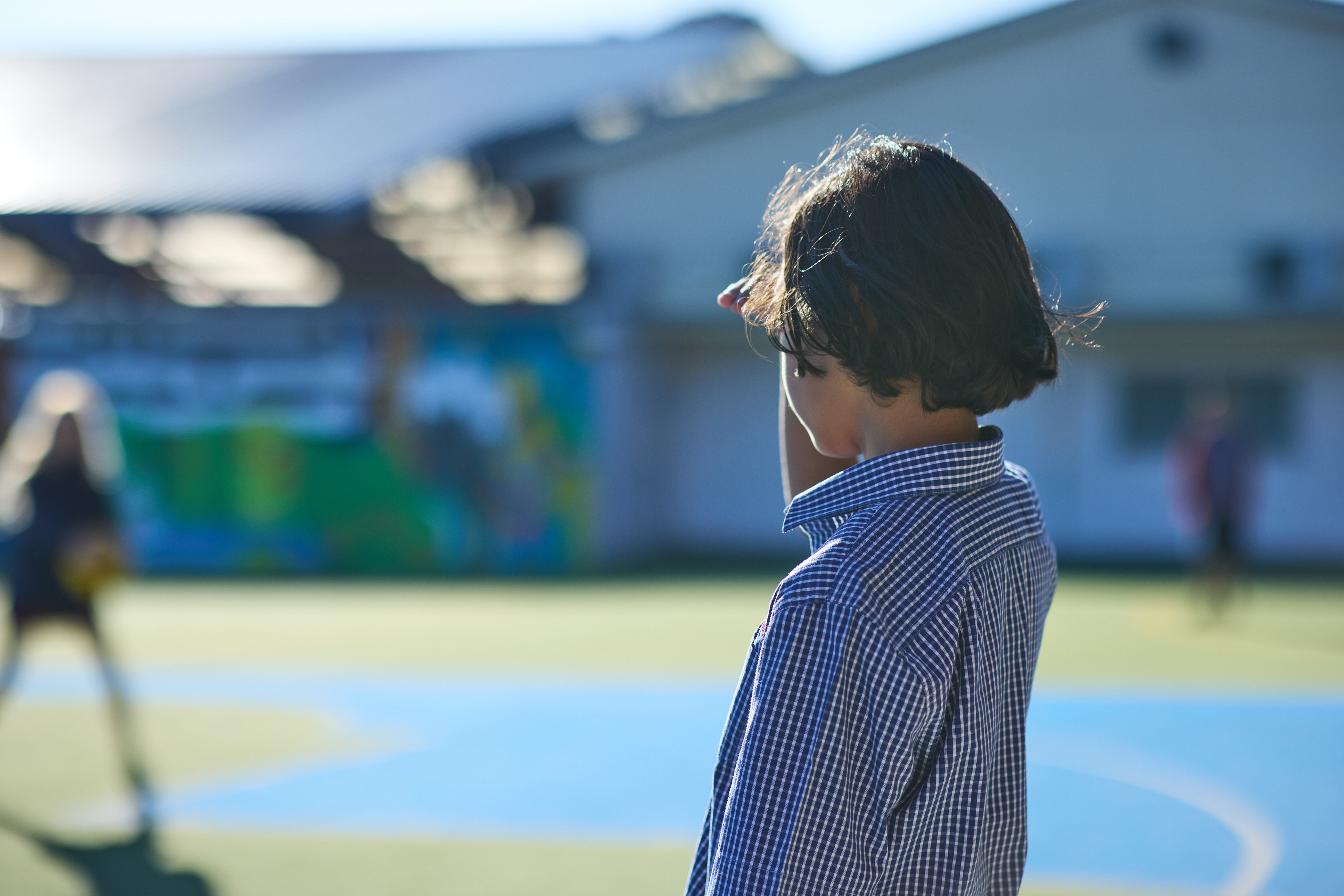 KidsCan child in a checkered shirt stands on a sunny playground, shielding their eyes. Blurred background of a building and other children playing.